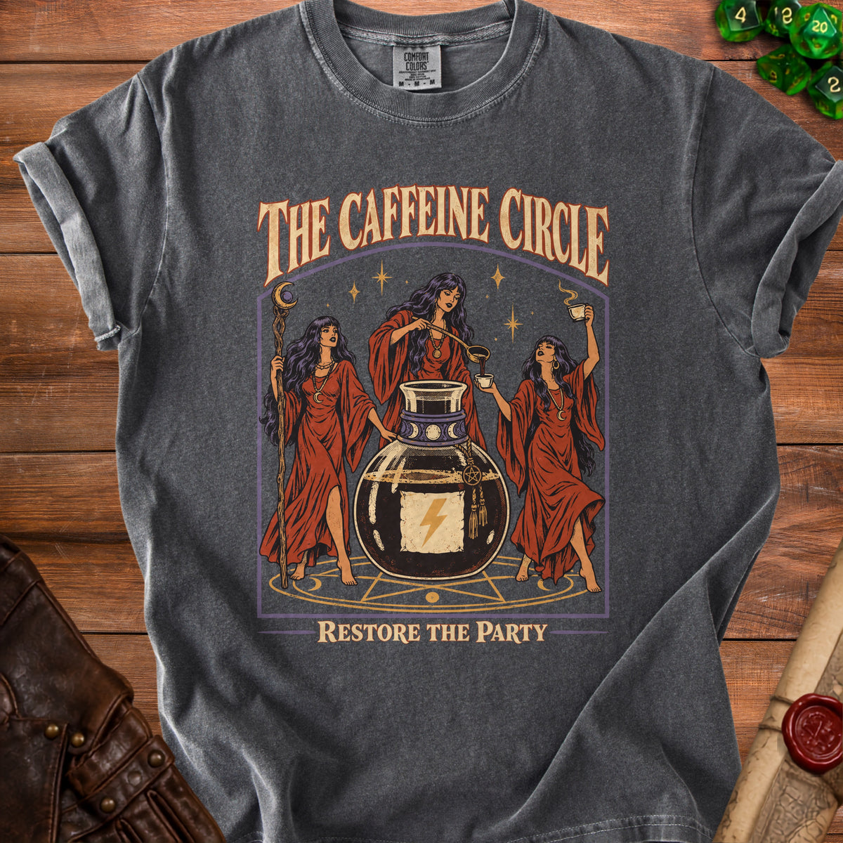 Dark grey t-shirt graphic with three witches around a potion bottle, text "THE CAFFEINE CIRCLE RESTORE THE PARTY".