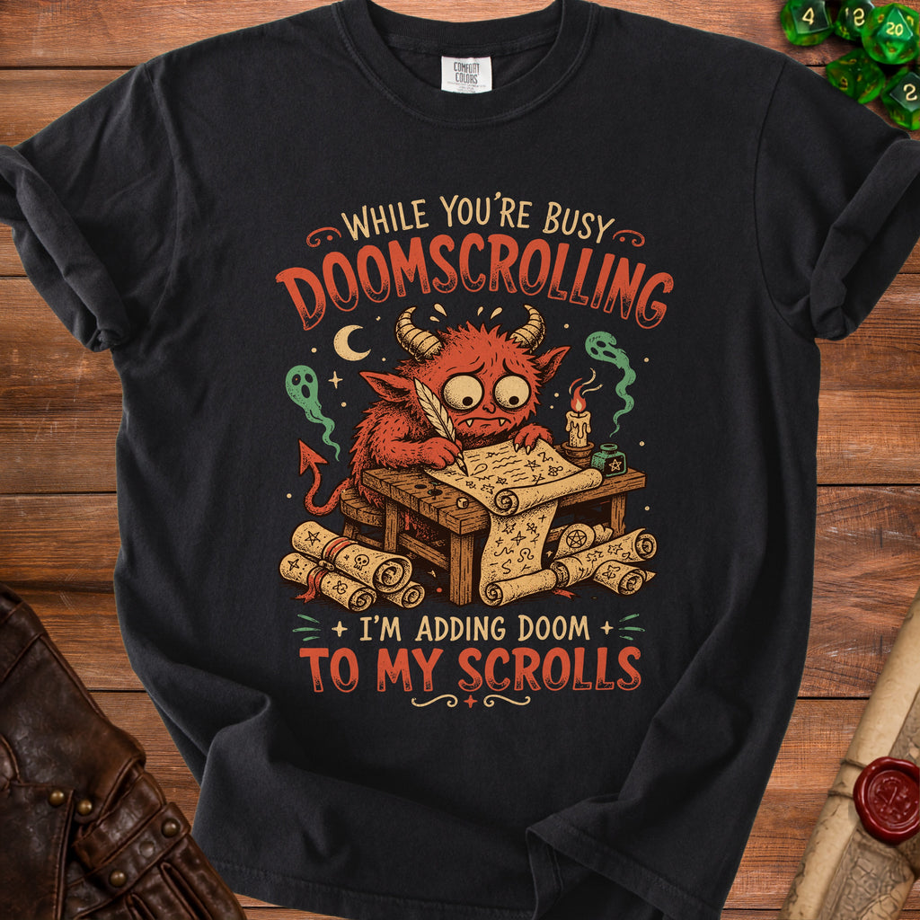 Black t-shirt with a cartoon demon writing scrolls, reading "While You're Busy Doomscrolling, I'm Adding Doom to My Scrolls".
