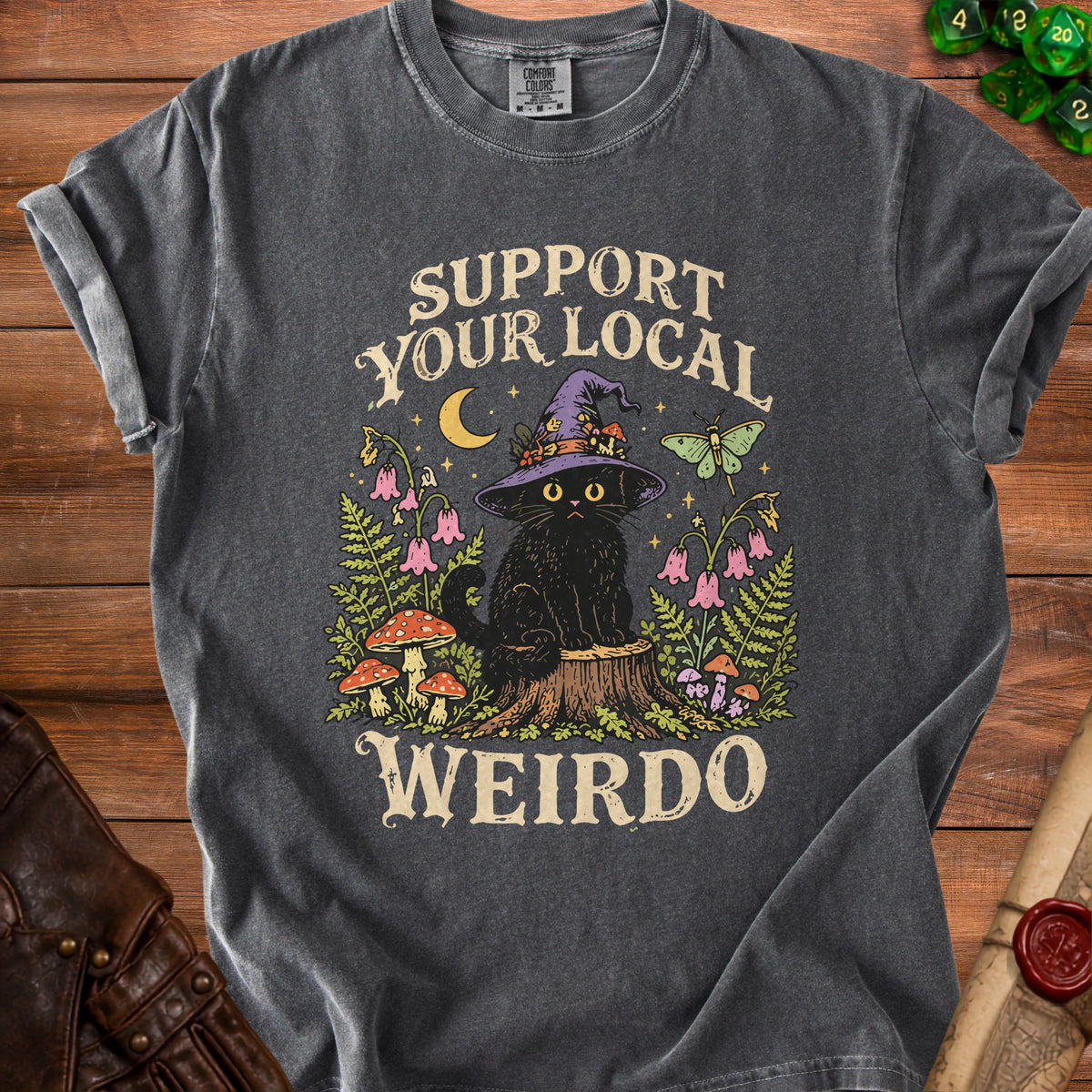 A dark gray t-shirt featuring a graphic of a black cat wearing a witch hat, surrounded by nature and the text SUPPORT YOUR LOCAL WEIRDO.