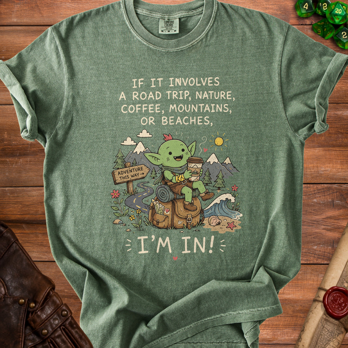Green t-shirt with a goblin character, travel theme, and text: "IF IT INVOLVES A ROAD TRIP, NATURE, COFFEE, MOUNTAINS, OR BEACHES, I'M IN!"
