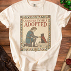 Cream t-shirt with medieval-style art of a person offering a cape to a black cat.