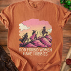 Rust-colored t-shirt featuring three fantasy adventurers climbing a rocky hill beneath the text GOD FORBID WOMEN HAVE HOBBIES.