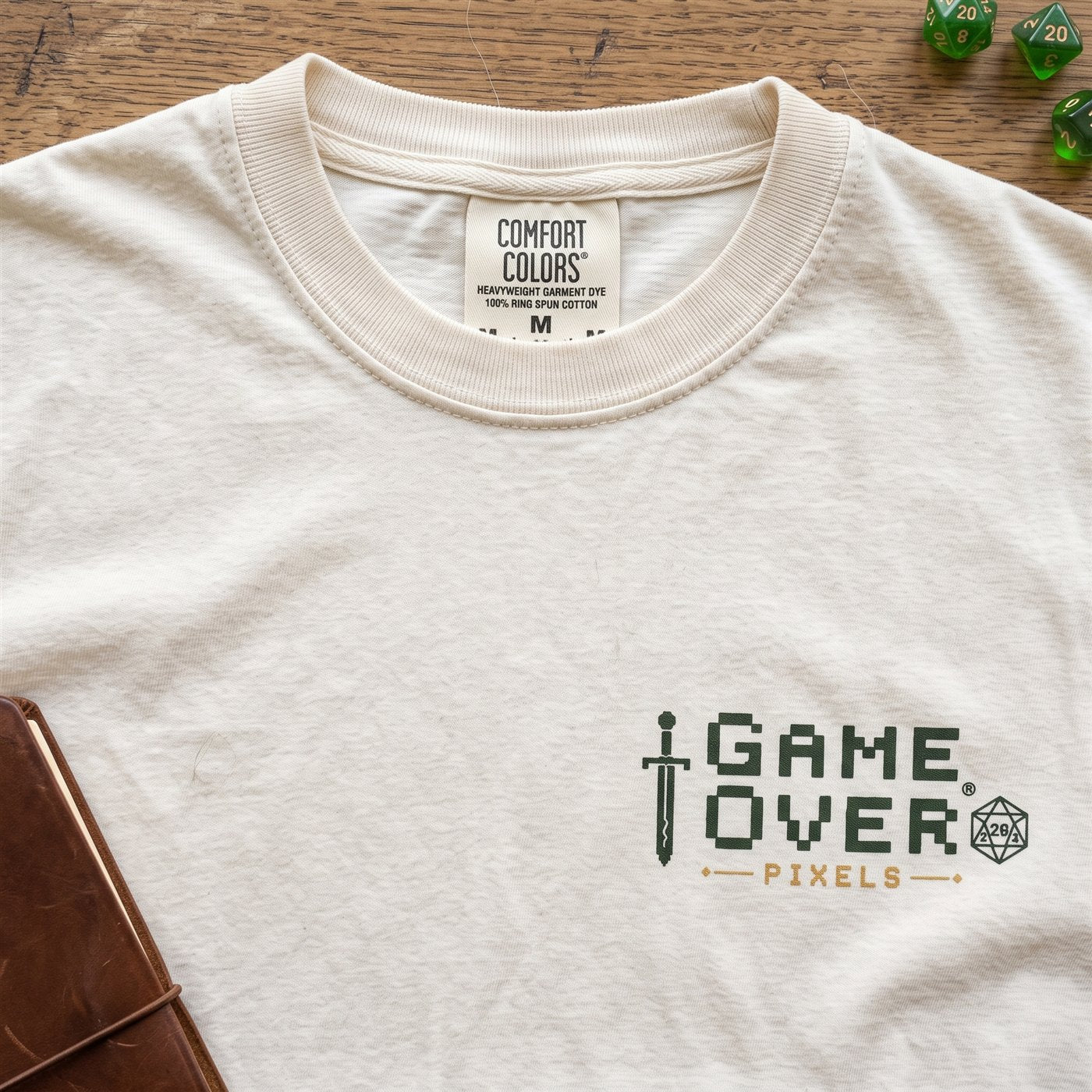 The Against My Will and Frog Wizard adventure tees on a cabin window seat beside a packed satchel, dice and a map